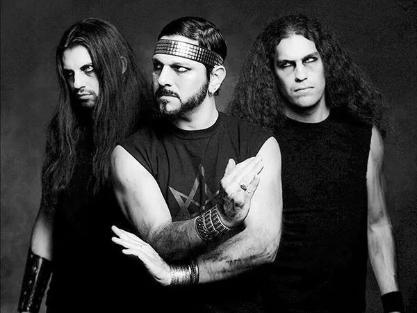 Three men with long dark hair and serious expressions pose in black sleeveless outfits against a dark background, giving a dramatic and intense vibe. One wears a studded headband and metallic wristbands.