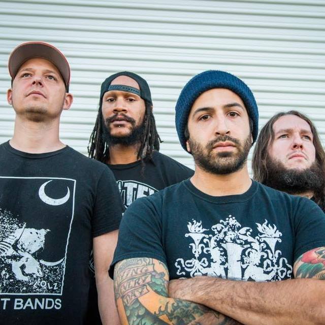 Four men stand in front of a white corrugated metal wall, looking serious. The man in front has tattooed arms and wears a blue beanie; the others wear t-shirts and caps, with neutral or focused expressions.