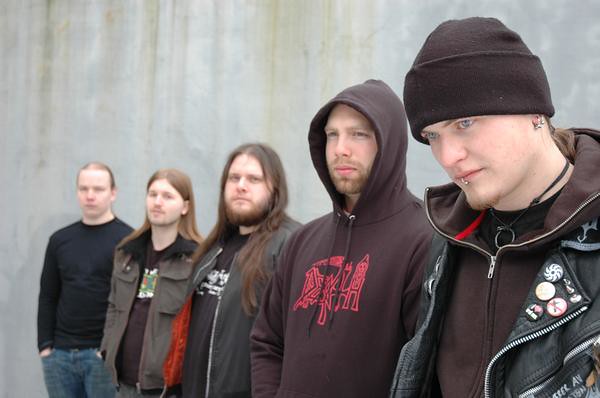 Five men with long hair and beards, dressed in dark clothing and hoodies, stand in a line against a gray, weathered wall. The man in front wears a beanie, jacket, and has facial piercings.