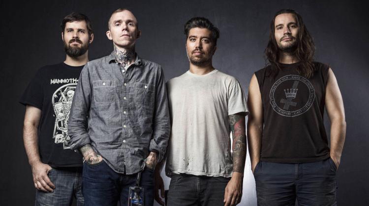 Four men stand side by side against a dark background. They have serious expressions and visible tattoos. Their clothing is casual, including T-shirts and a button-up shirt.