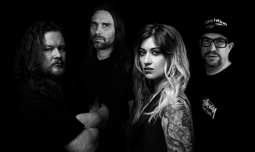 Four people pose together against a black background; three men and one woman with long hair and a tattooed arm, all dressed in dark clothing, looking seriously at the camera.