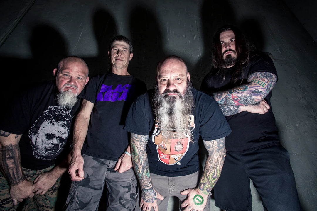 Four men with tattoos, beards, and intense expressions stand close together looking down at the camera. They wear dark t-shirts and shorts, with a concrete wall behind them in dramatic lighting.