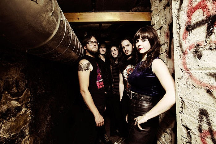 Five people dressed in black stand among tall, flowering plants in a greenhouse, looking at the camera with serious expressions. Bright red flowers surround them on both sides.